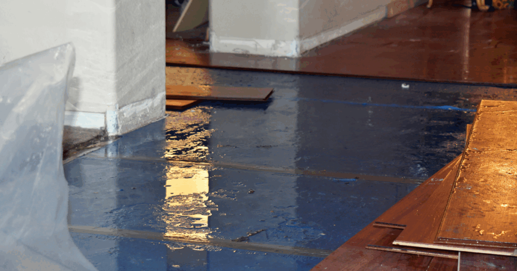 A room with water on the floor, partially removed wooden flooring, and visible water damage on the walls, indicating a flood or significant water leak.